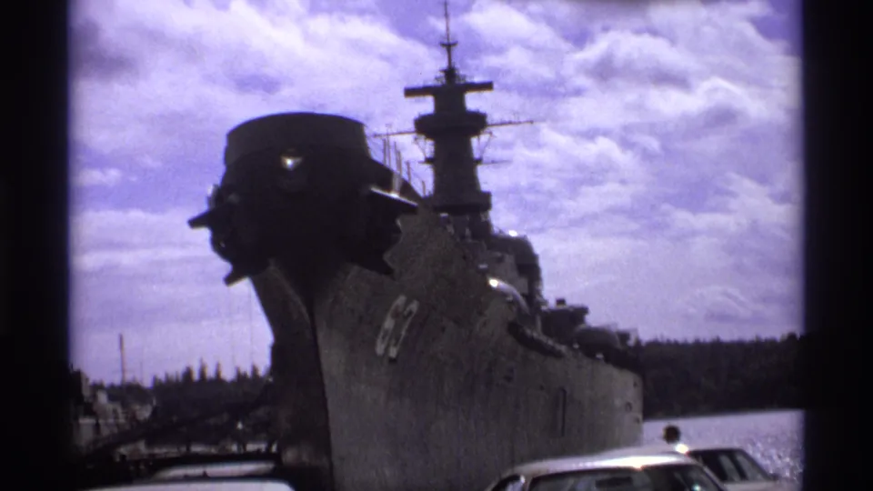 Closer 1974 Vancouver frame of a gray military ship bow, deck structures, mast, cloudy sky, and shoreline water.