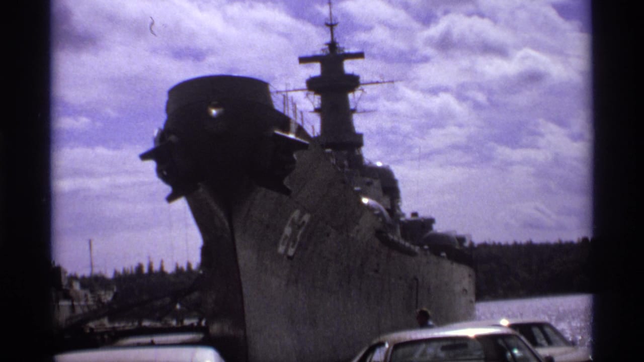 Gray naval ship bow marked 63 beside docked cars under a cloudy sky.