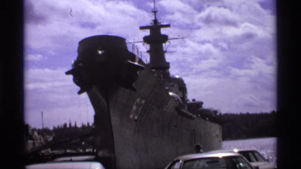 1974 Vancouver harbor view of a large gray naval vessel with bow number 63, dockside cars, water, trees, and cloudy sky.