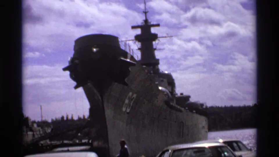 Dockside 8mm view in Vancouver of a large gray naval ship, its mast and hull dominating the frame near harbor water and parked cars.