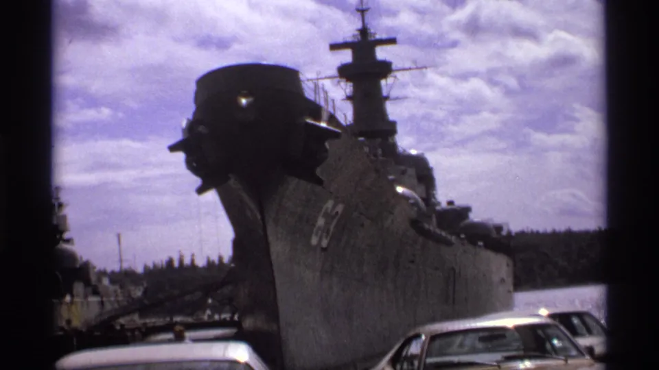 1974 Vancouver harbor footage showing the gray bow of a naval vessel marked 63, with parked 1970s cars and a wooded shore beyond.
