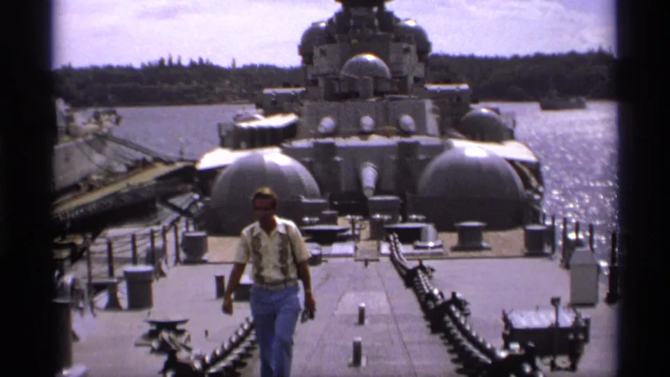 A man in a light shirt and jeans walks across the deck of a large naval vessel in Vancouver, with turrets, railings, and harbor water visible.