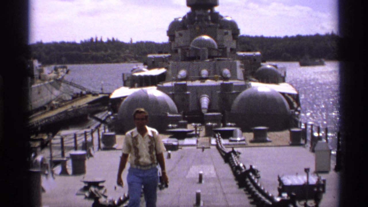 Man walking on the deck of a gray historic ship with turrets, water, and shoreline behind him.