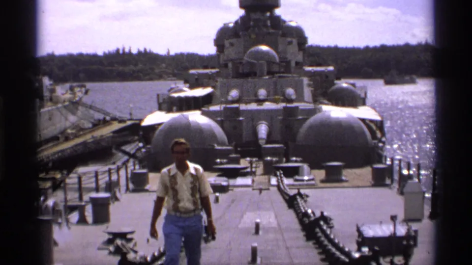 1974 Vancouver home-movie frame of a man walking across a gray warship deck with large turrets, water, and wooded shore in view.