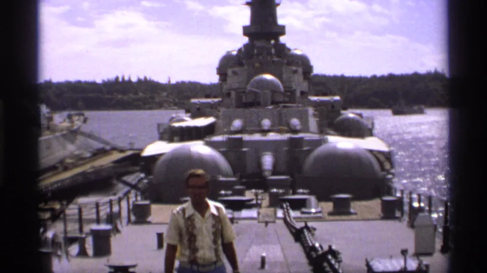 A man appears near the foreground of a historic ship deck in Vancouver, with gray naval structures and water behind him.