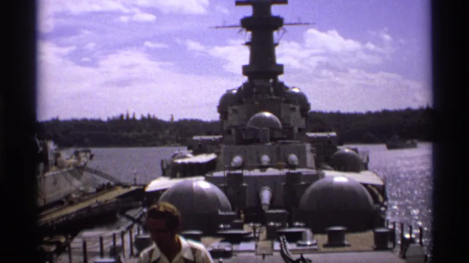 1970s 8mm frame looking along a gray warship deck toward its superstructure, with a visitor partly cut off in the foreground.