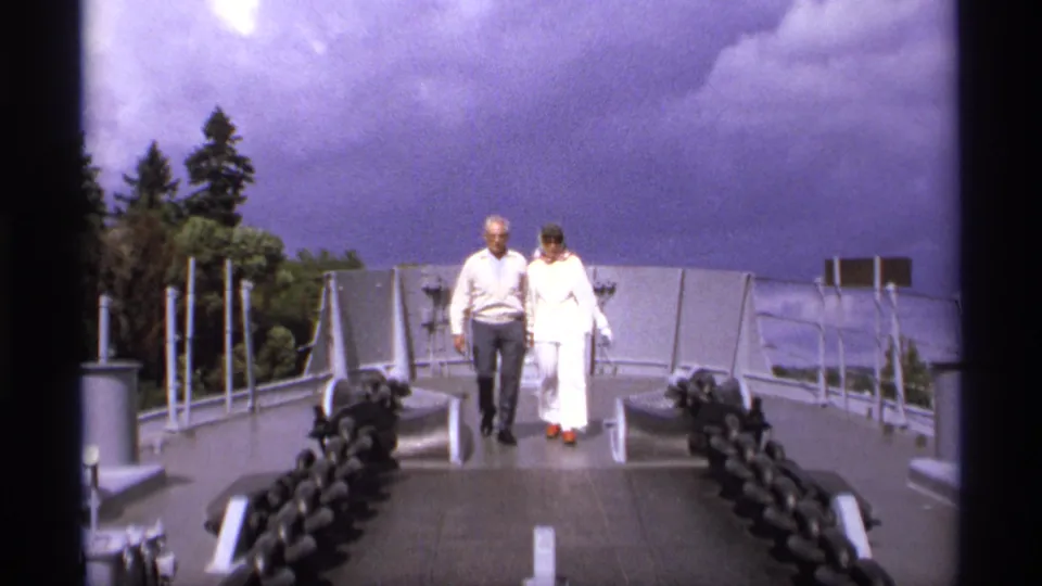 An older couple walks along a gray naval ship deck in 1974 Vancouver footage, framed by railings, deck fittings, and a dramatic clouded sky.