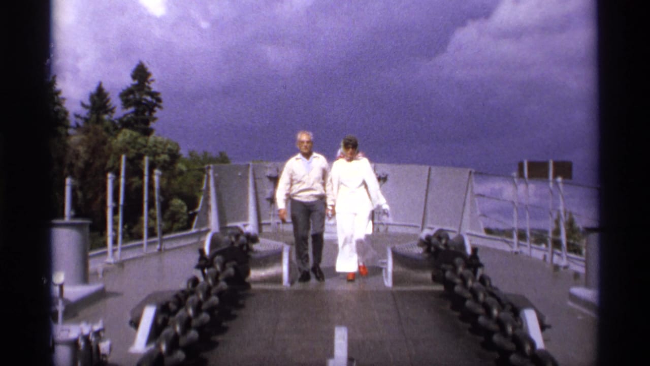 Elderly couple walking hand-in-hand on a bridge walkway with chain railings.