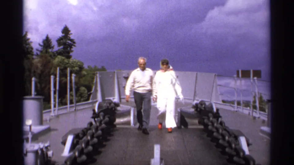The couple continues toward the camera on the Vancouver bridge walkway, framed by chains, railings and trees.