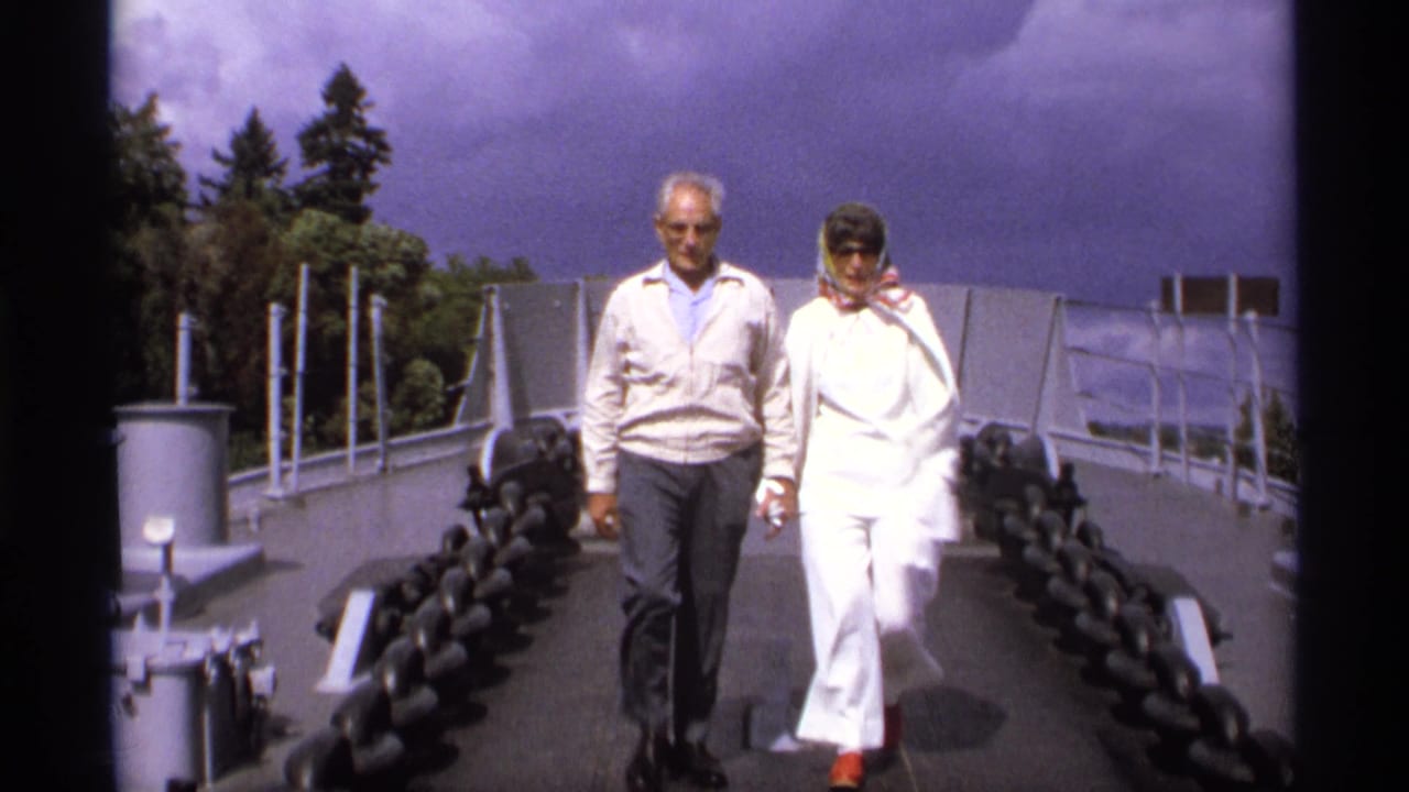 Two adults walk along a battleship deck between heavy chains and railings.