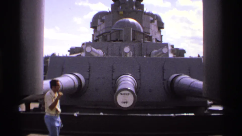 The visitor pauses near the gray battleship turret, with several large barrels and the ship structure visible behind him.