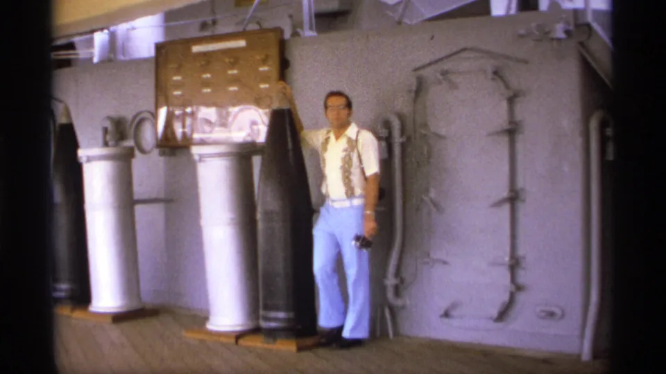 Inside or beside a ship display area, the man stands next to tall artillery shells, a large casing, and a heavy gray hatch.