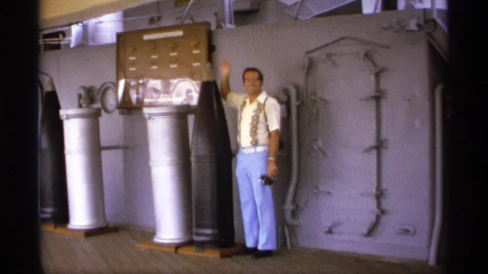 The man waves toward the camera while standing beside shipboard ordnance, a display case, pipes, and a large hatch in the 1974 Vancouver footage.