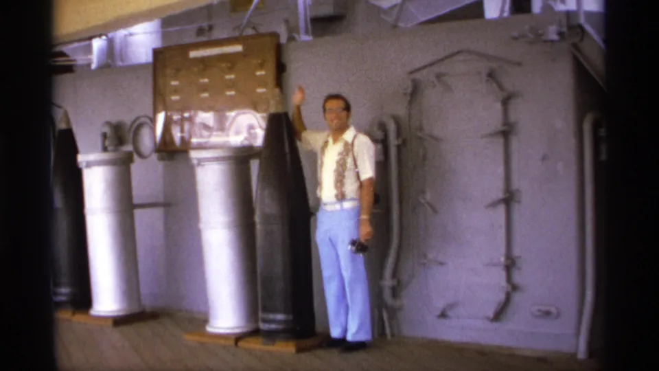A man holds one hand up beside large projectiles, silver canisters, a display board, and a hatch on the USS Missouri deck visit footage.