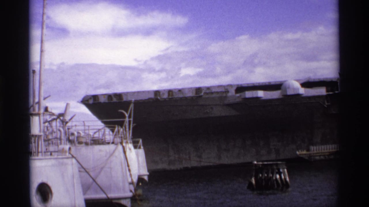 Large gray aircraft carrier seen across harbor water under a cloudy sky.