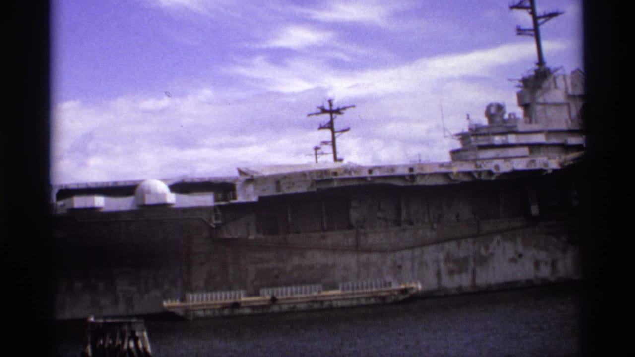 Aircraft carrier side with island superstructure and radar domes beside harbor water.