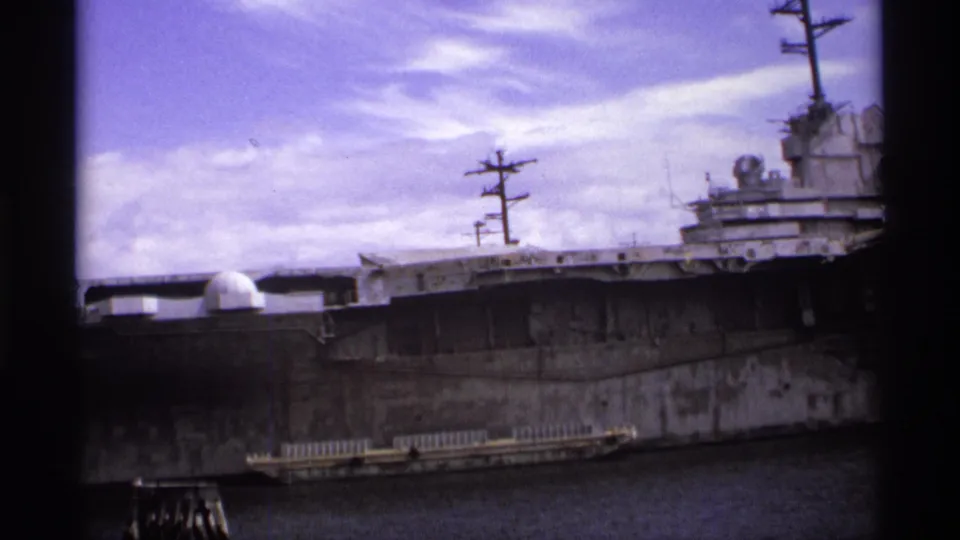 A docked aircraft carrier fills the frame, with its island, masts, radar domes, and long gray hull visible in Vancouver harbor.