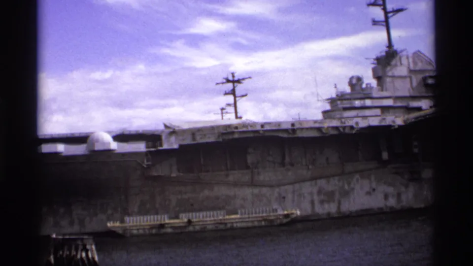 The frame centers on the carrier's long side, deck edge, mast silhouettes, and radar domes, with harbor water below and clouds above.