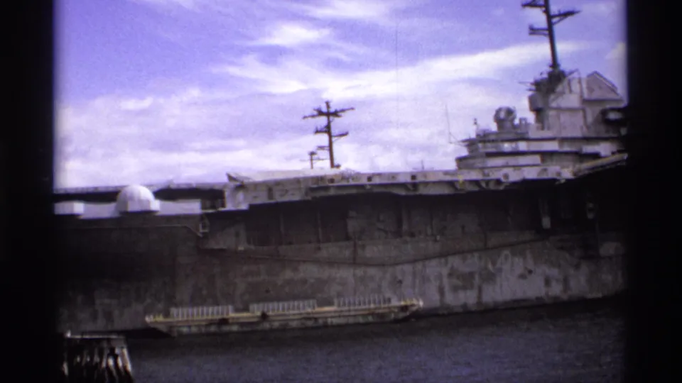 Final frame of the 8mm clip shows the docked naval carrier from the side, including its superstructure, masts, gray hull, and harbor water.