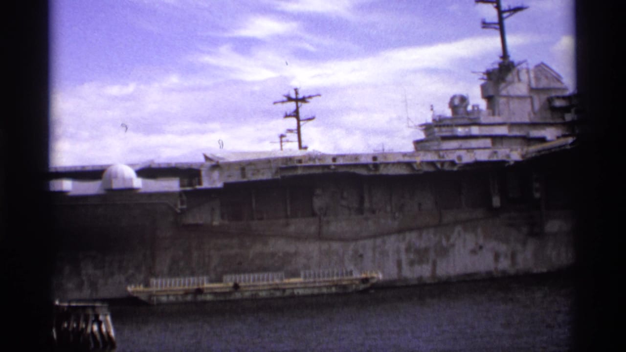 Large gray warship docked beside water, seen through dark vertical frame borders.