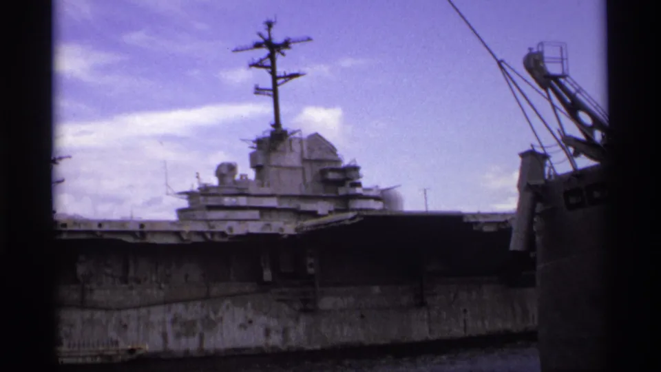 1974 Vancouver home-movie frame showing the warship superstructure, mast, dark hull, and nearby crane across the water.