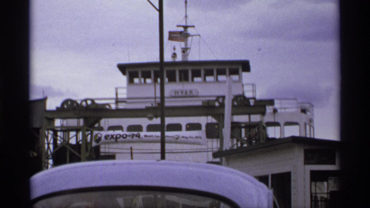 1974 home-movie frame showing the white HYAK ferry at a dock, partly blocked by a vehicle roof, with an Expo 74 banner visible.