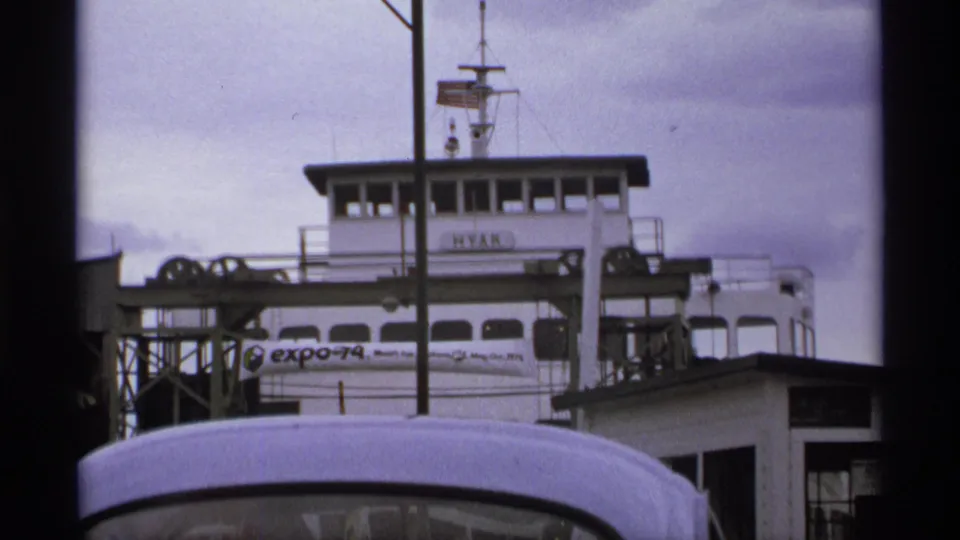 1974 home-movie frame showing the white HYAK ferry at a dock, partly blocked by a vehicle roof, with an Expo 74 banner visible.