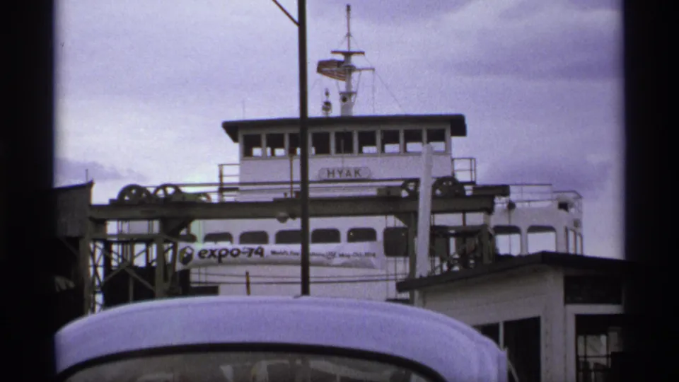 1974 home-movie still of the HYAK ferry at a dock, with Expo 74 signage and a vehicle roof blocking the lower frame.
