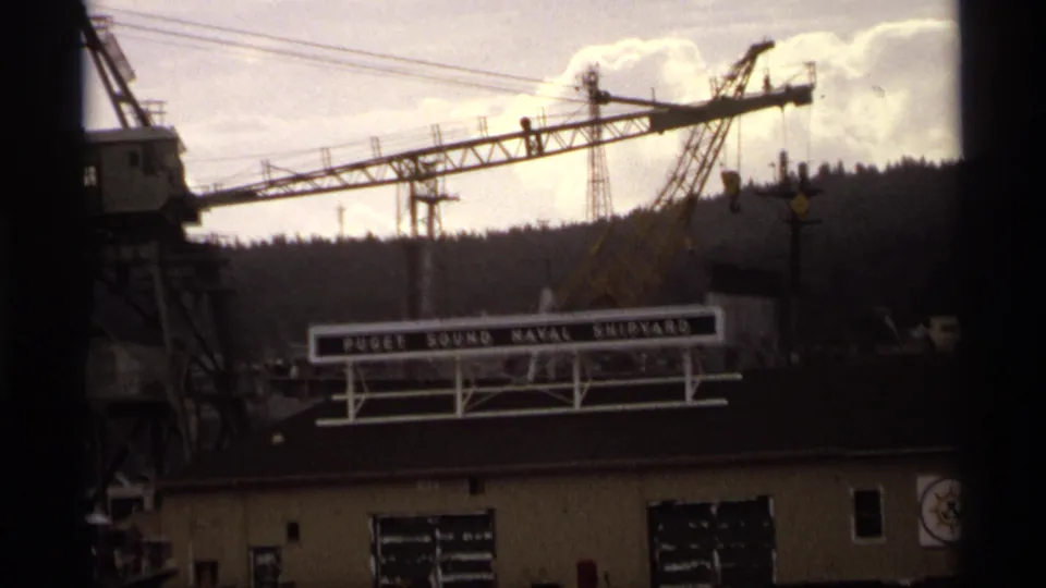 1974 home-movie frame showing a Puget Sound Naval Shipyard sign below large cranes, with wooded shoreline behind.