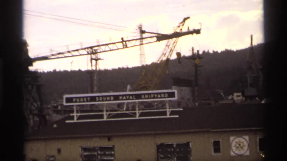 1974 8mm frame with the Puget Sound Naval Shipyard sign, crane booms, and industrial waterfront structures under a pale sky.
