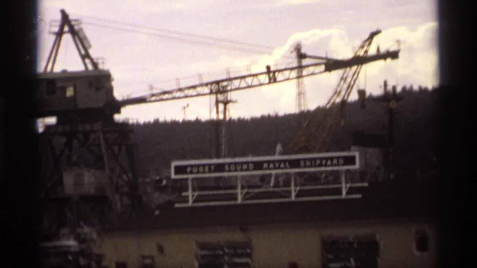 1974 8mm frame showing a crane tower at left, the Puget Sound Naval Shipyard sign, and industrial buildings along the waterfront.