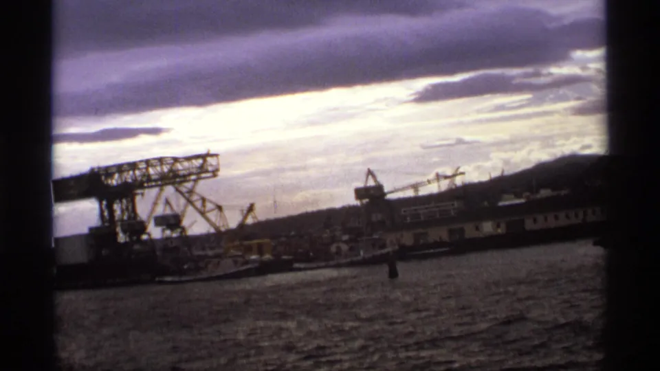 Wide 1974 8mm harbor view with shipyard cranes, waterfront buildings, open water, and a dramatic cloudy sky.