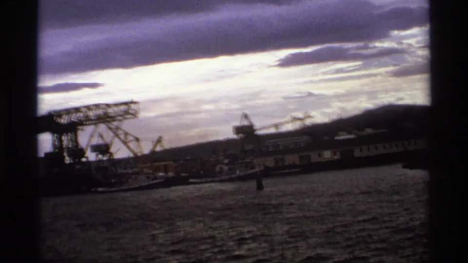 Vancouver industrial waterfront in 1974, showing cranes, a long vessel, low hills, dark water, and bright clouded sky.