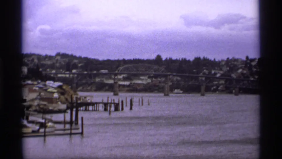 1974 Vancouver river view with a steel bridge, docked boats, wooden pilings, and houses along the far shore.