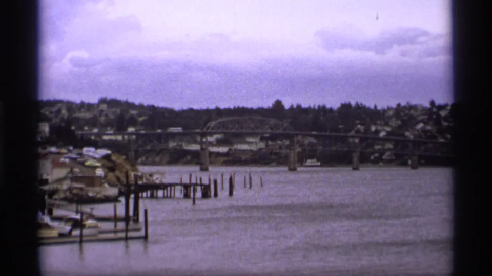 1974 Vancouver waterfront frame with docks and pilings in the foreground, a bridge across the river, and homes on the far bank.
