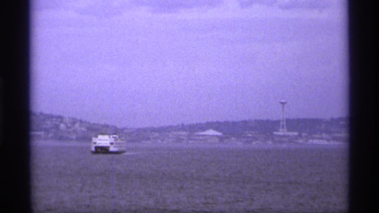 1974 Vancouver home-movie frame showing a small white ferry crossing hazy water toward a distant shoreline and needle-like tower.