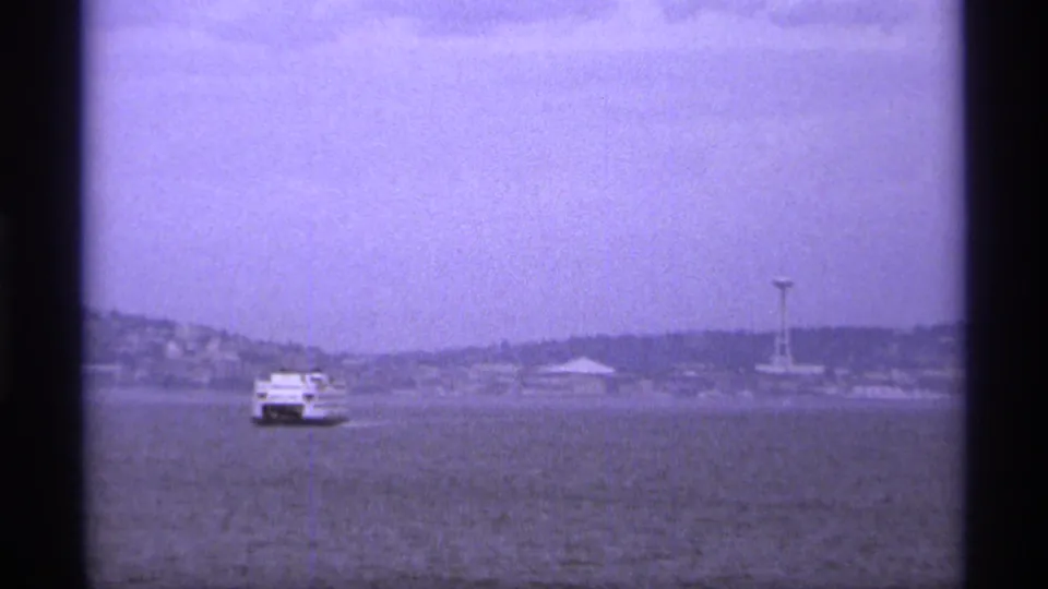 1974 Vancouver home-movie frame showing a small white ferry crossing hazy water toward a distant shoreline and needle-like tower.
