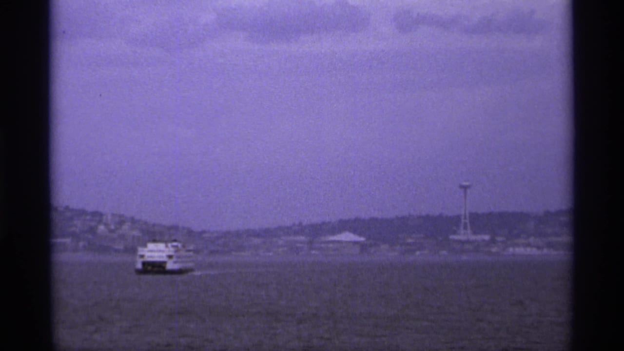 Small vessel on gray-blue water with shoreline buildings and a distant tower.