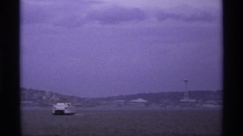 A purple-blue tinted 8mm frame shows the boat low in the scene, with distant hills, shoreline structures, and a tower through haze.