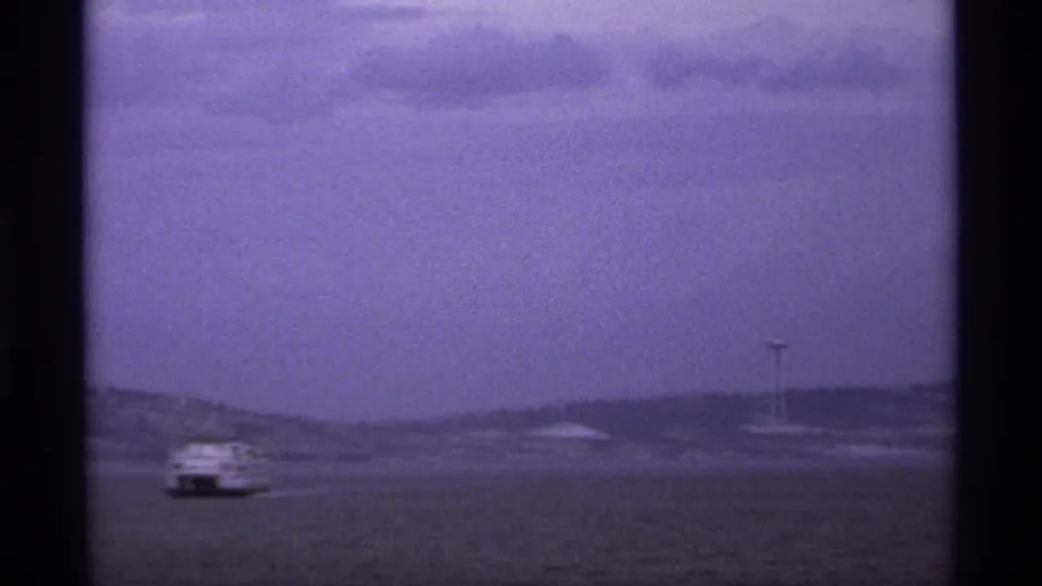 Final 1974 waterfront frame with a small boat crossing the bay, low hills on the horizon, and a distant needle-like tower.