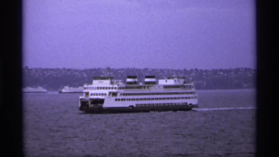 A large white ferry moves across Vancouver water in 1974, with a wooded or built-up shoreline behind it under a purple-tinged sky.