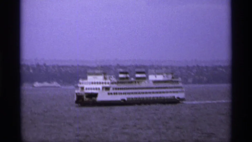1974 8mm-style waterfront frame showing a white ferry moving across water near a distant shoreline.