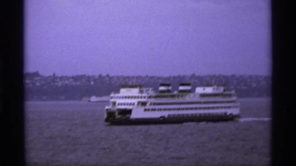 A white multi-deck ferry continues across the waterfront in a grainy 1974 8mm home-movie frame.