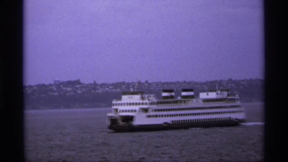 Grainy 1974 waterfront view of a white ferry passing across open water with shoreline structures beyond.
