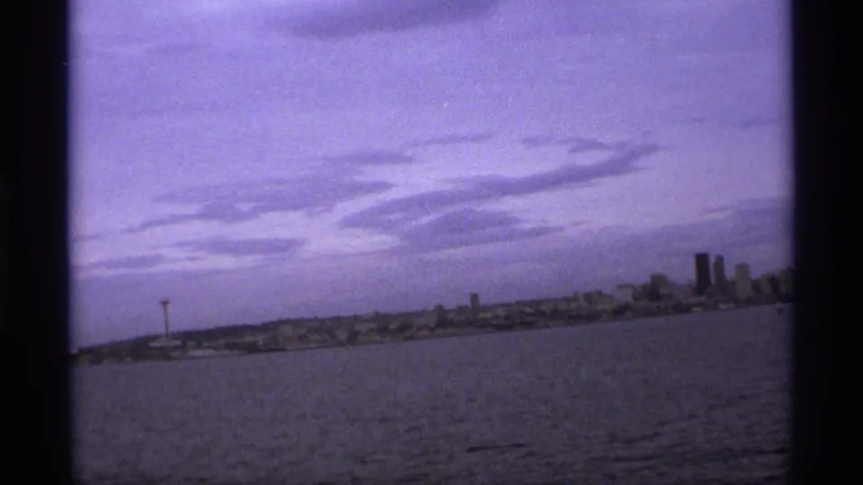 1974 8mm-style view across water toward a distant skyline with a needle-like observation tower and taller buildings.