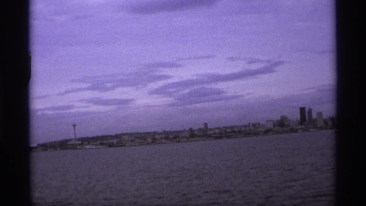 1974 8mm waterfront skyline view, with water in the foreground, a distant observation tower, clustered buildings, and a purple sky.