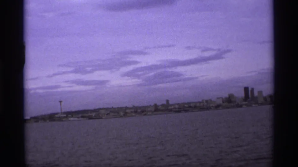 1974 8mm waterfront skyline view, with water in the foreground, a distant observation tower, clustered buildings, and a purple sky.