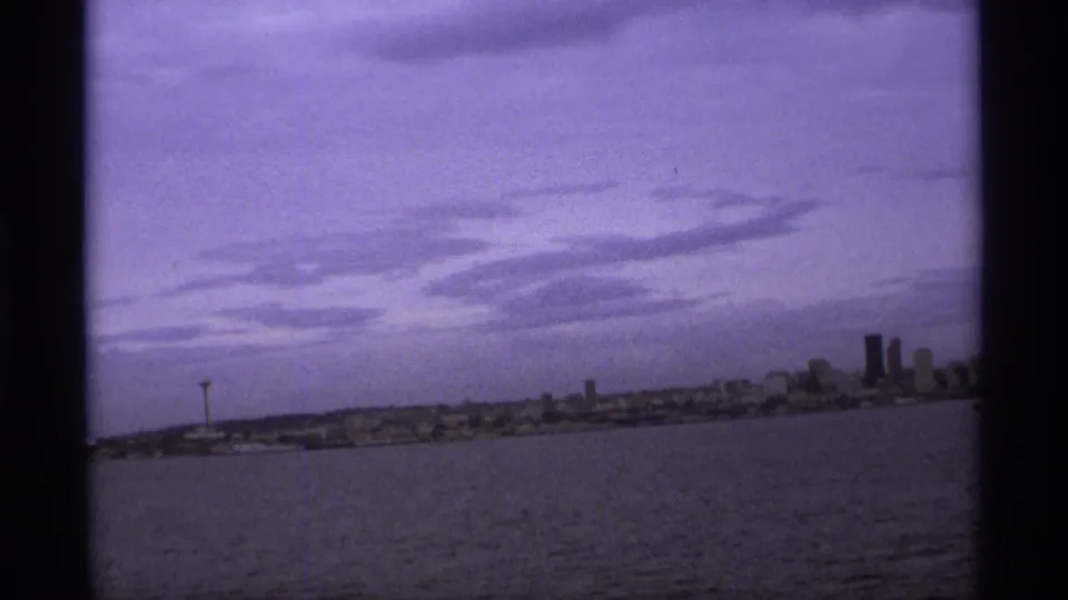 Handheld 1974 8mm view across water toward a city skyline, with an observation tower left of center and taller buildings at right.