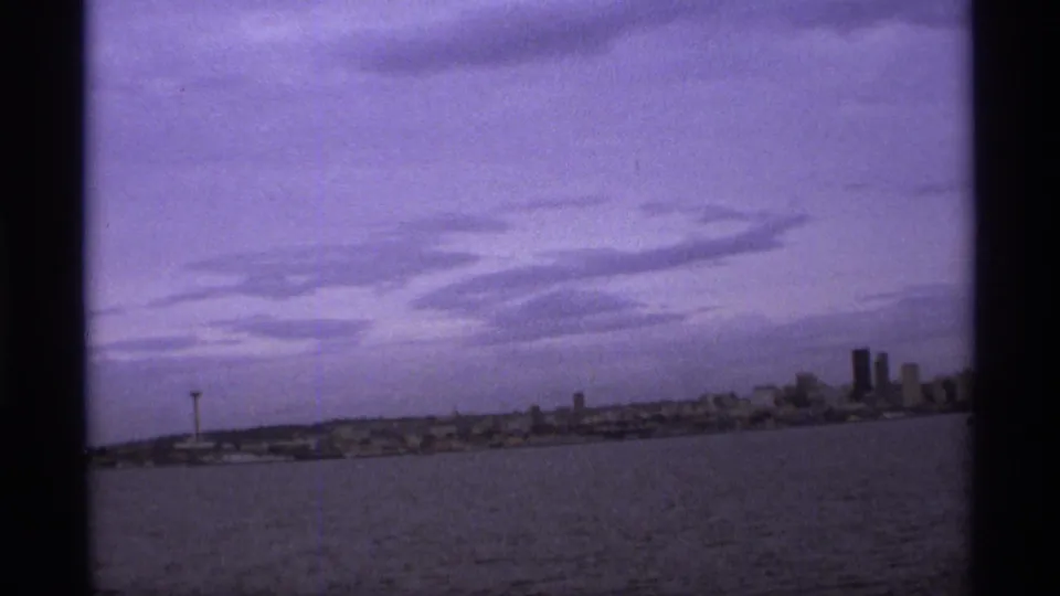 1974 8mm panoramic waterfront frame showing a low skyline, observation tower, distant high-rises, water, and soft cloud bands.