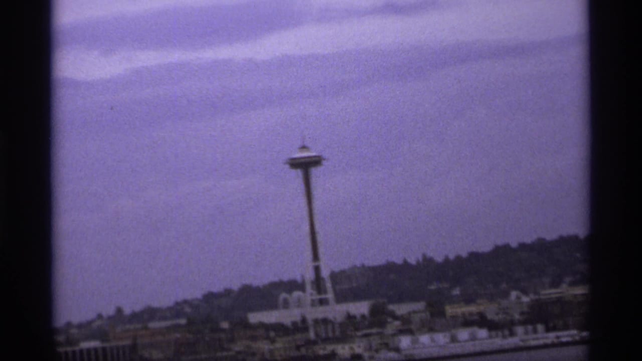 1970s 8mm frame showing the Space Needle from across water, with low buildings and wooded hills below a muted sky.